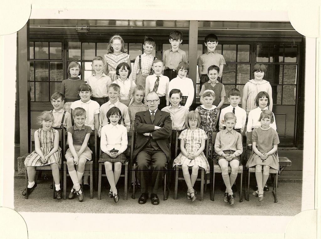 Class of about twenty-seven junior-school children in four rows, an older male teacher in glasses seated in the centre with his arms folded, in front of glazed classroom doors.