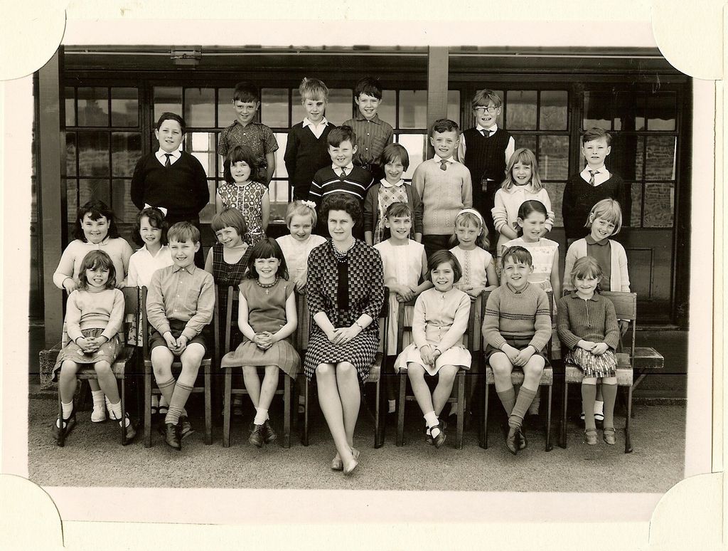 Class of about thirty-three children in four rows, a woman teacher in a checked dress seated in the centre of the front row, in front of a veranda with glazed doors.