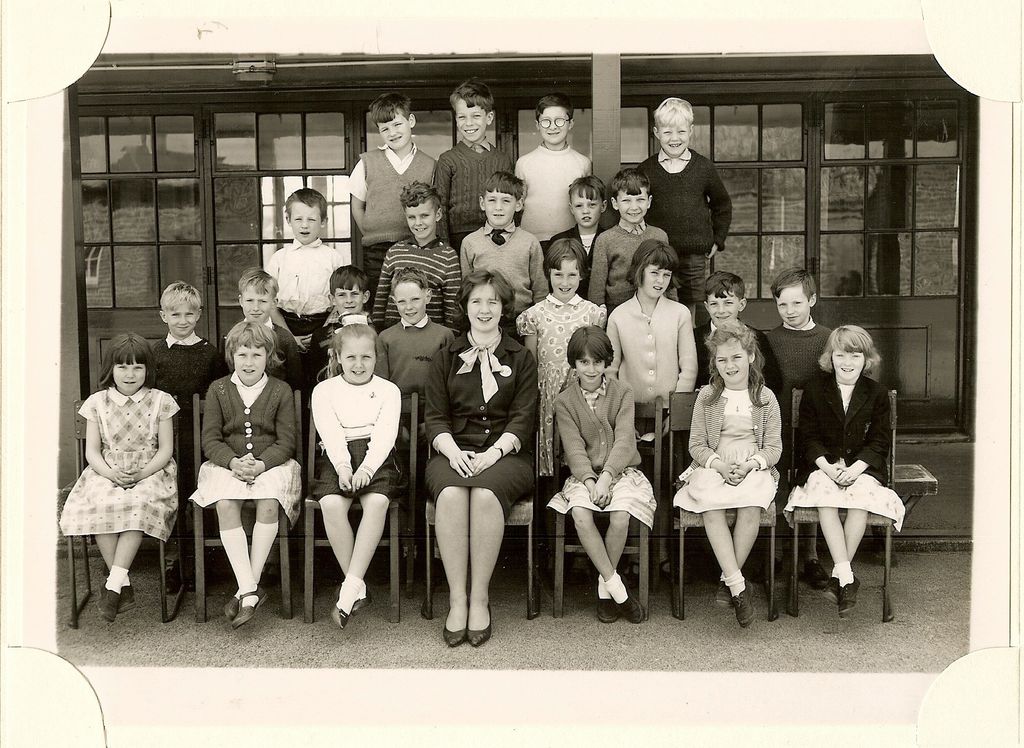 Class of about thirty-three children in four rows, a woman teacher in a dark dress with a bow at the neck seated in the centre of the front row.