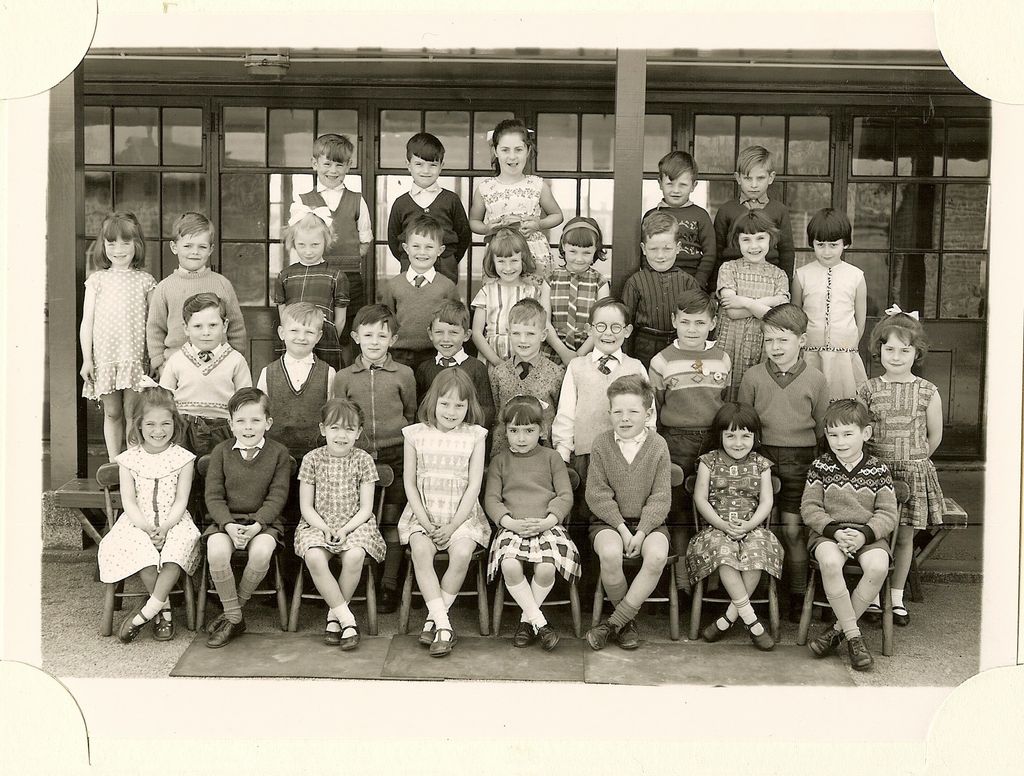 Class of about thirty-eight infants in four rows, the front row on small chairs set on mats, in front of a veranda with glazed doors; no teacher appears.