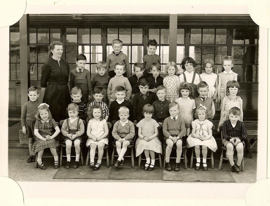 Class of about thirty-five infants in four rows, a woman teacher in a dark dress standing at the left, in front of glazed classroom doors.