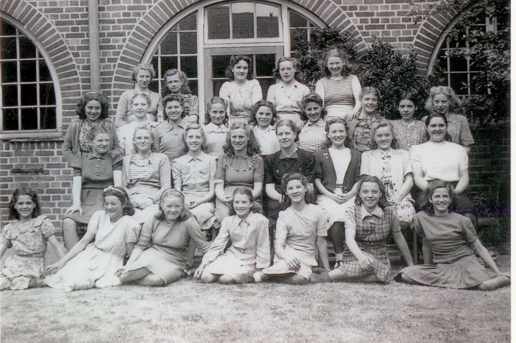 About thirty teenage girls in summer dresses and blouses in four rows, the front row sitting on the grass, in front of a brick school building with large arched windows.