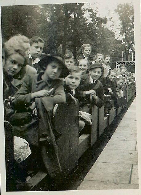 Children in coats and caps, some in glasses and school hats, lean out of the open wooden carriages of a miniature railway, looking at the camera, with trees behind.