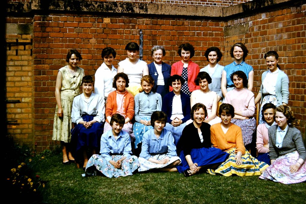Colour photo of about twenty teenage girls in cardigans and full skirts in three rows on a lawn in the corner of a brick building, with an older woman teacher in a blue suit in the centre of the back row.