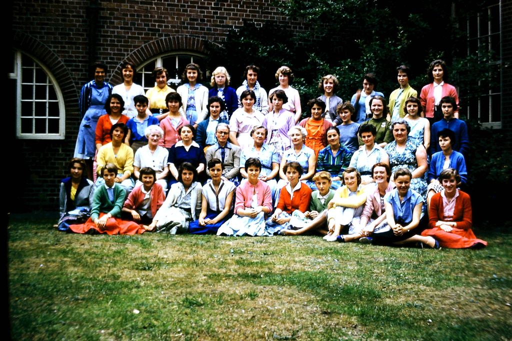 Colour photo of about fifty girls and women teachers in five rows on a lawn in front of a brick school building with an arched window; the girls wear bright cardigans and skirts.