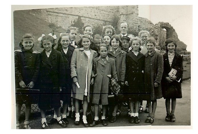About twenty girls in winter coats and white socks, some with hair ribbons, stand in a group with a male teacher in front of the ruined stone walls of Dudley Castle.