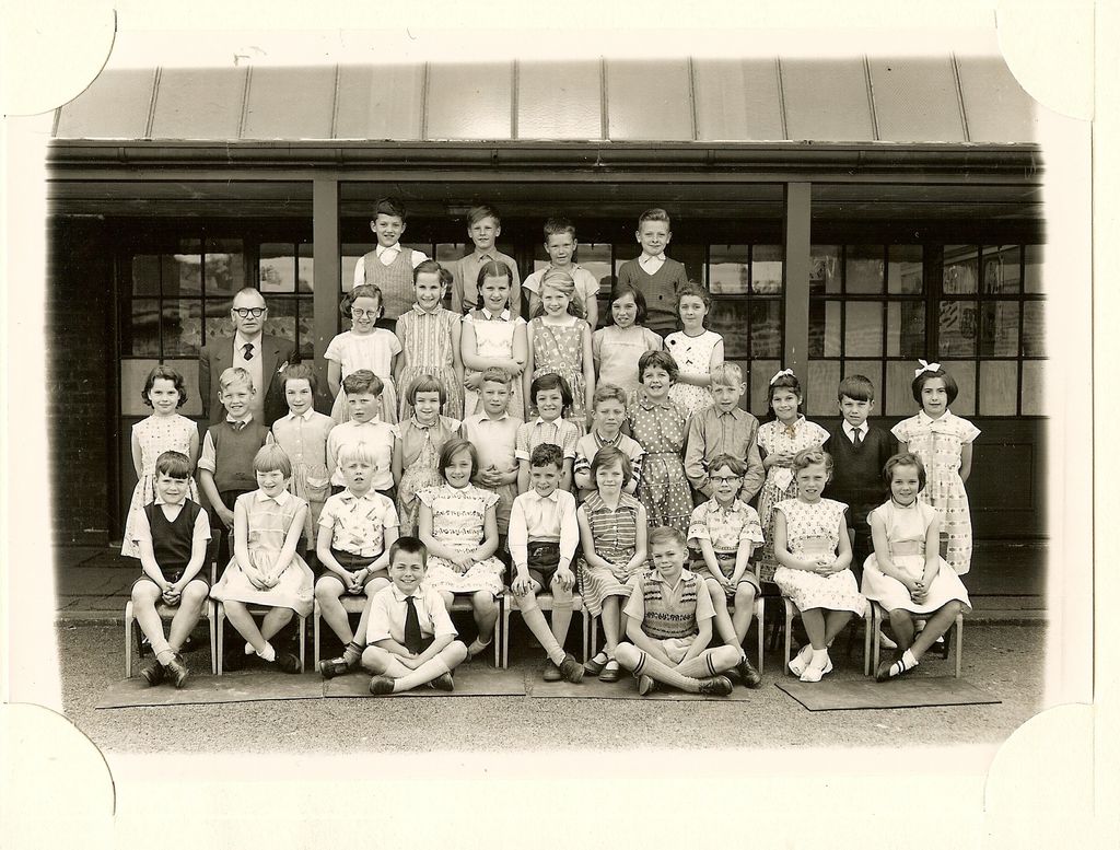 Large class of about forty-five junior-school children in five rows on the playground, a male teacher in glasses standing at the left, in front of a veranda with glazed doors under a pitched canopy.