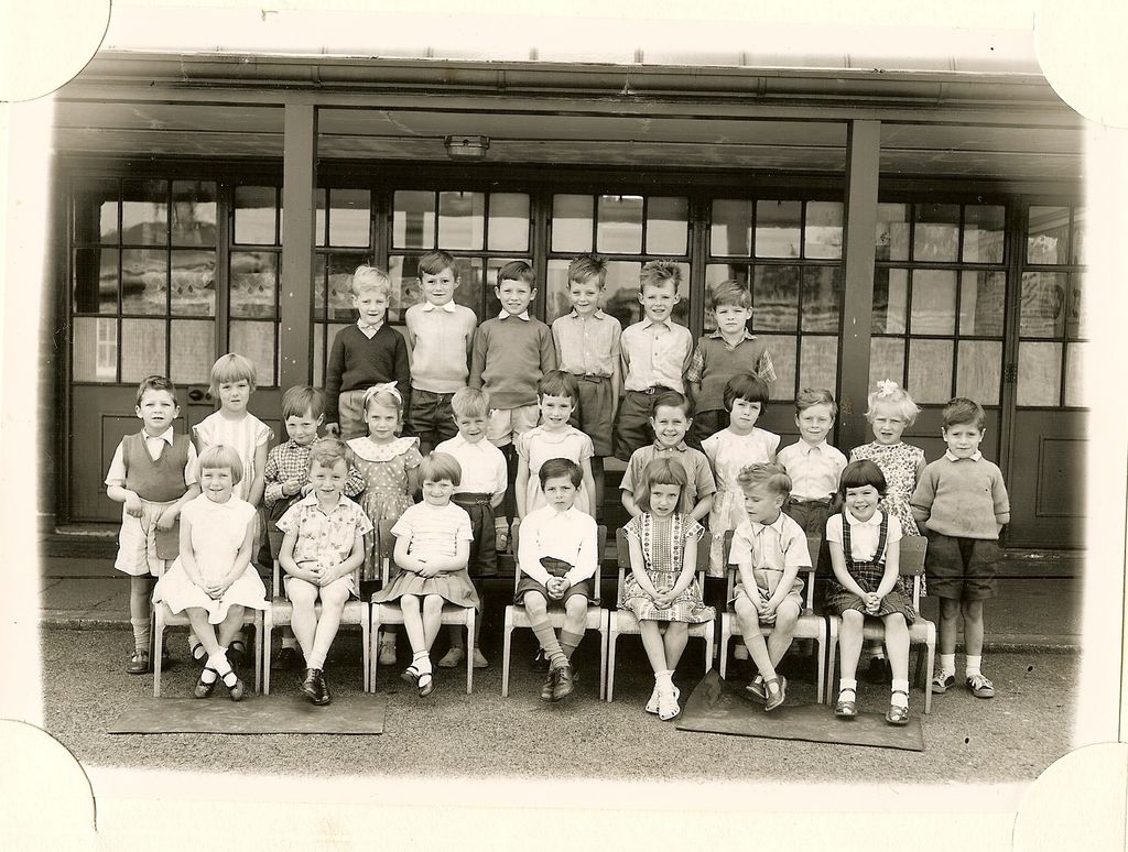 Class of about thirty infants in four rows, the front row on small chairs on mats, in front of a veranda with glazed doors; no teacher appears.