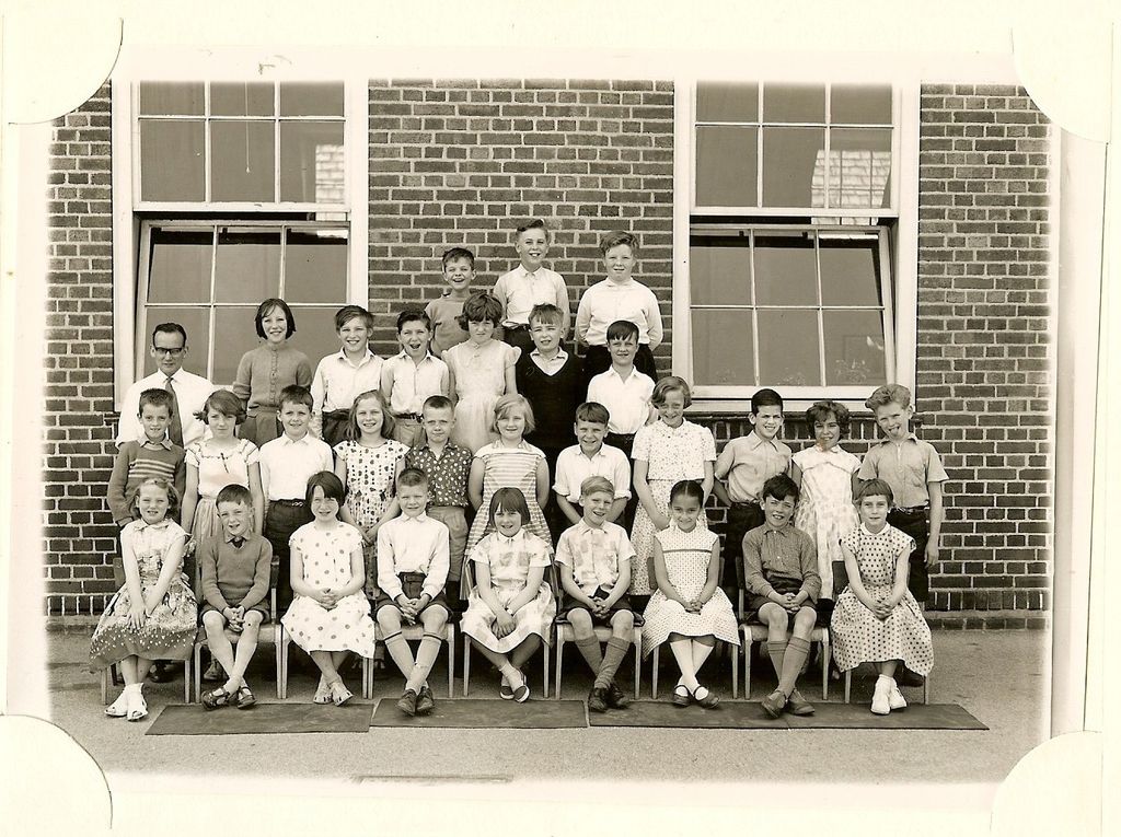 Class of about forty children in five rows in front of a brick school building with tall sash windows, a young male teacher in glasses standing at the left.