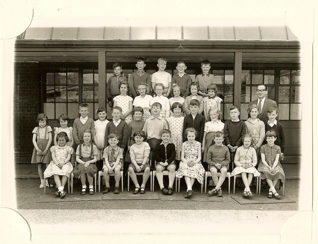 Class of about forty-five children in five rows, a male teacher in glasses standing at the right, in front of a veranda with glazed doors.