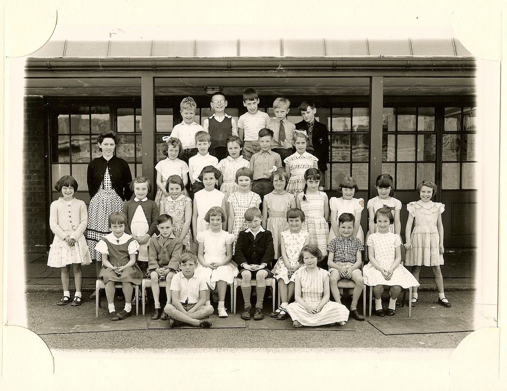 Class of about forty infants in five rows, a woman teacher in a dark cardigan standing at the left, with two children sitting on the ground in front.