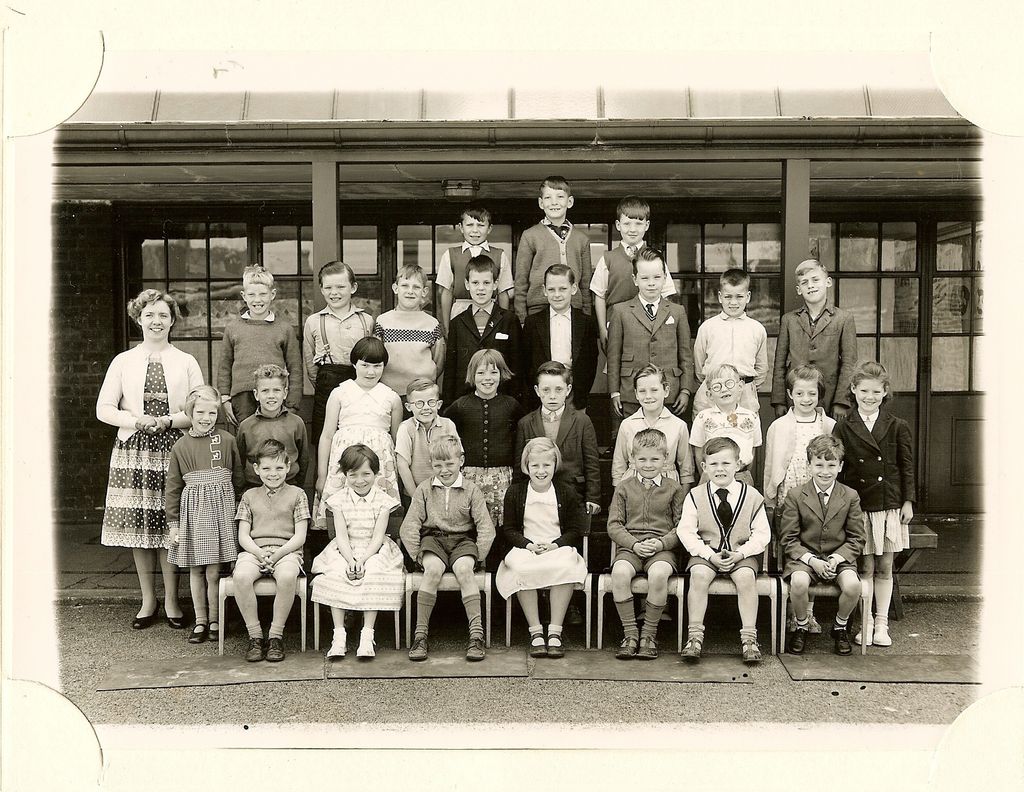 Class of about thirty-five children in five rows, a woman teacher in a light cardigan and patterned skirt standing at the left, in front of glazed classroom doors.