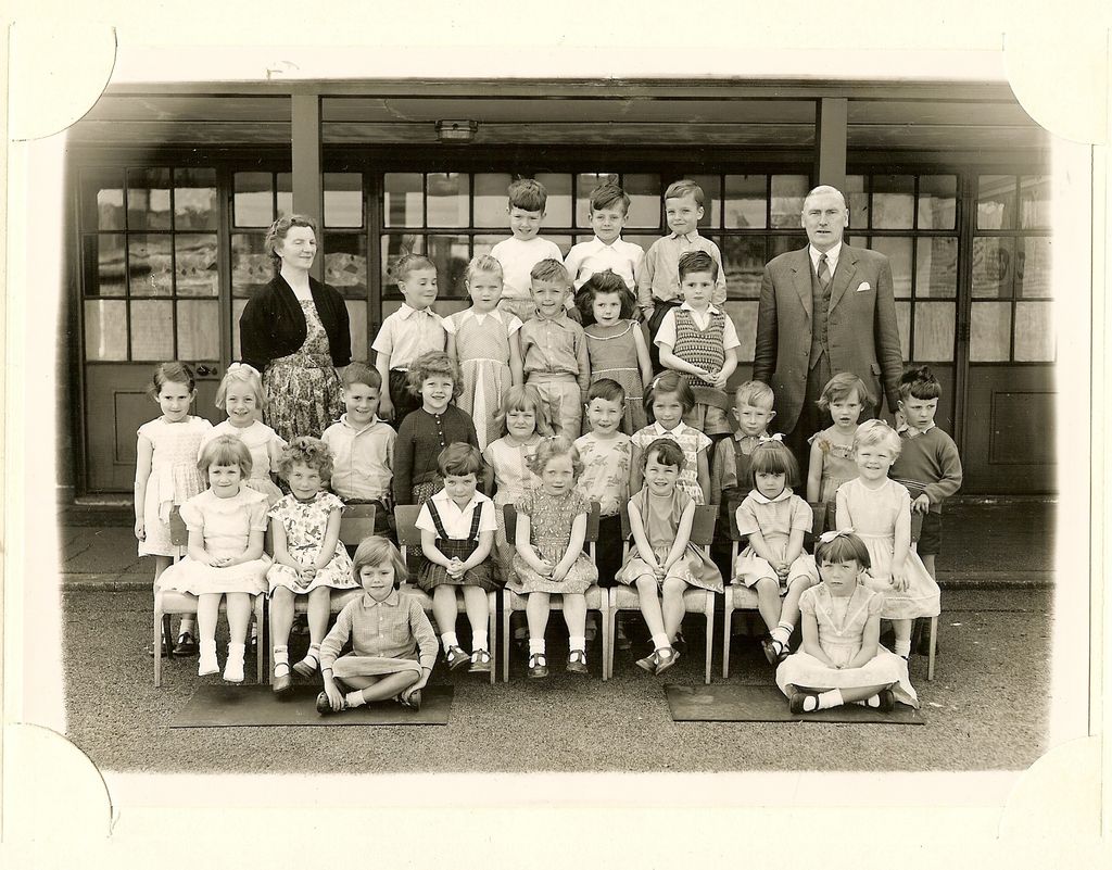 Class of about thirty-five infants in five rows, a woman teacher in a dark cardigan and an older man in a suit standing at the back, two girls sitting on the ground in front.