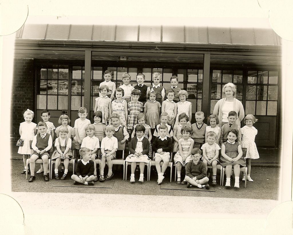 Class of about forty-five infants in five rows, an older woman teacher in a cardigan standing at the right, two boys sitting cross-legged in front.