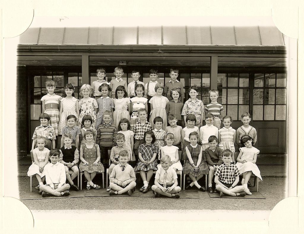 Class of about forty-five children in five rows, four boys sitting cross-legged in front, in front of glazed classroom doors; no teacher appears.