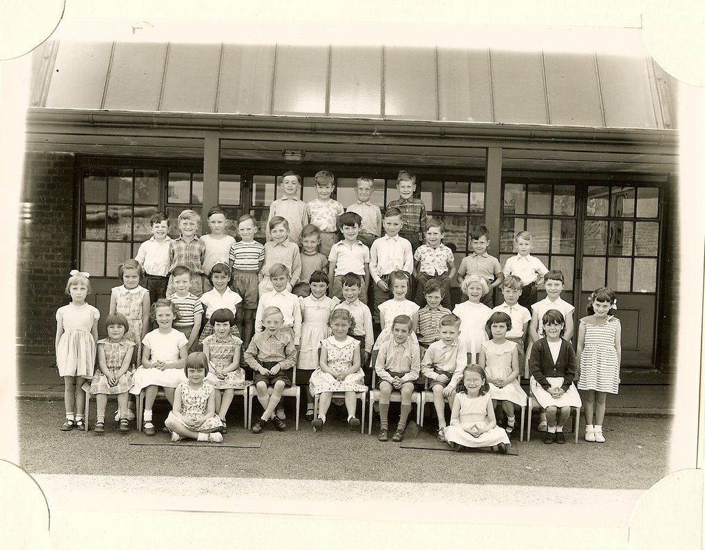 Class of about fifty young children in six rows under a veranda canopy, two girls sitting on the ground in front; no teacher appears.
