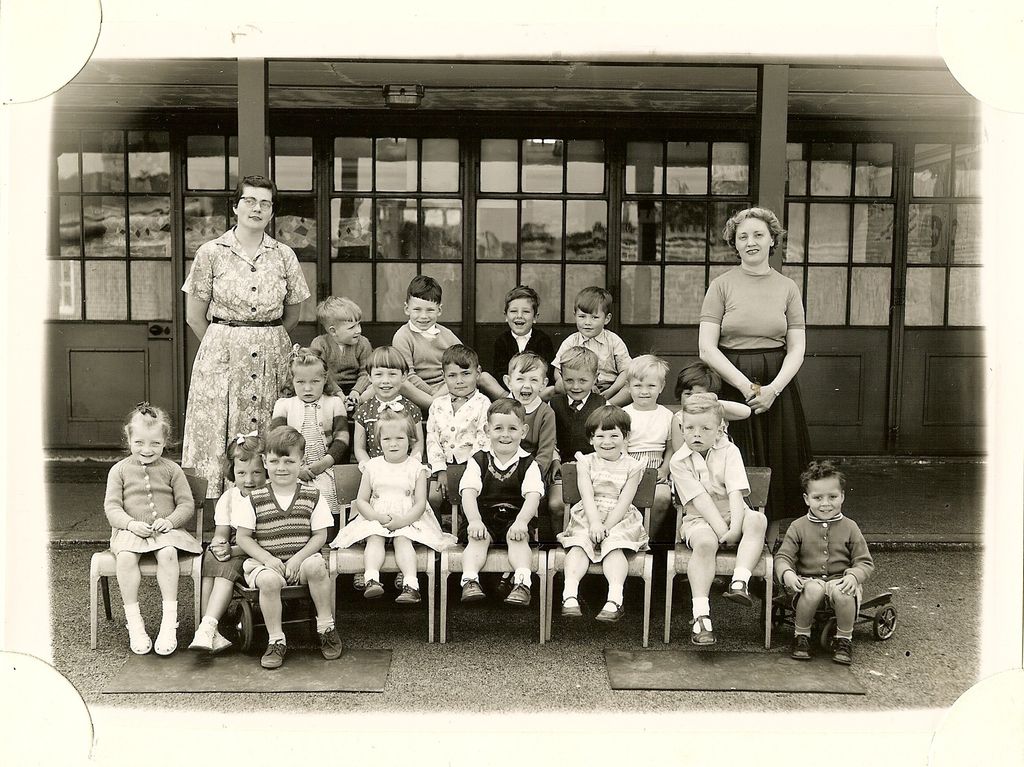 Small group of about twenty very young children with two women teachers, one boy sitting on a toy cart at the right, in front of glazed classroom doors.