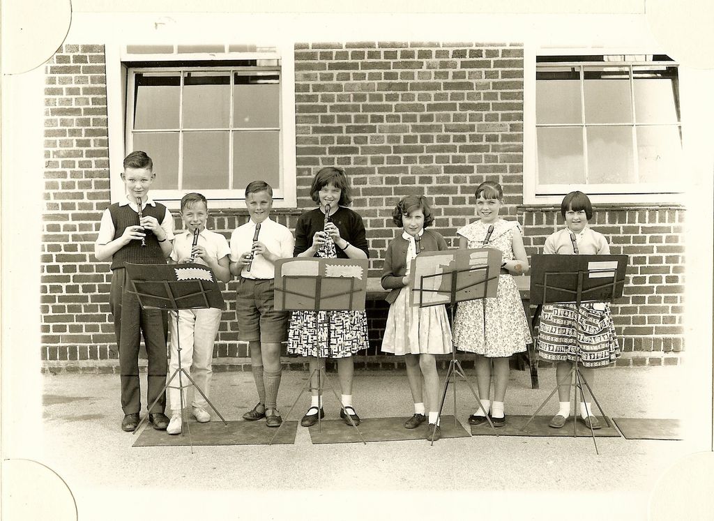 Seven children playing descant recorders at music stands in a row in front of a brick school wall with sash windows.