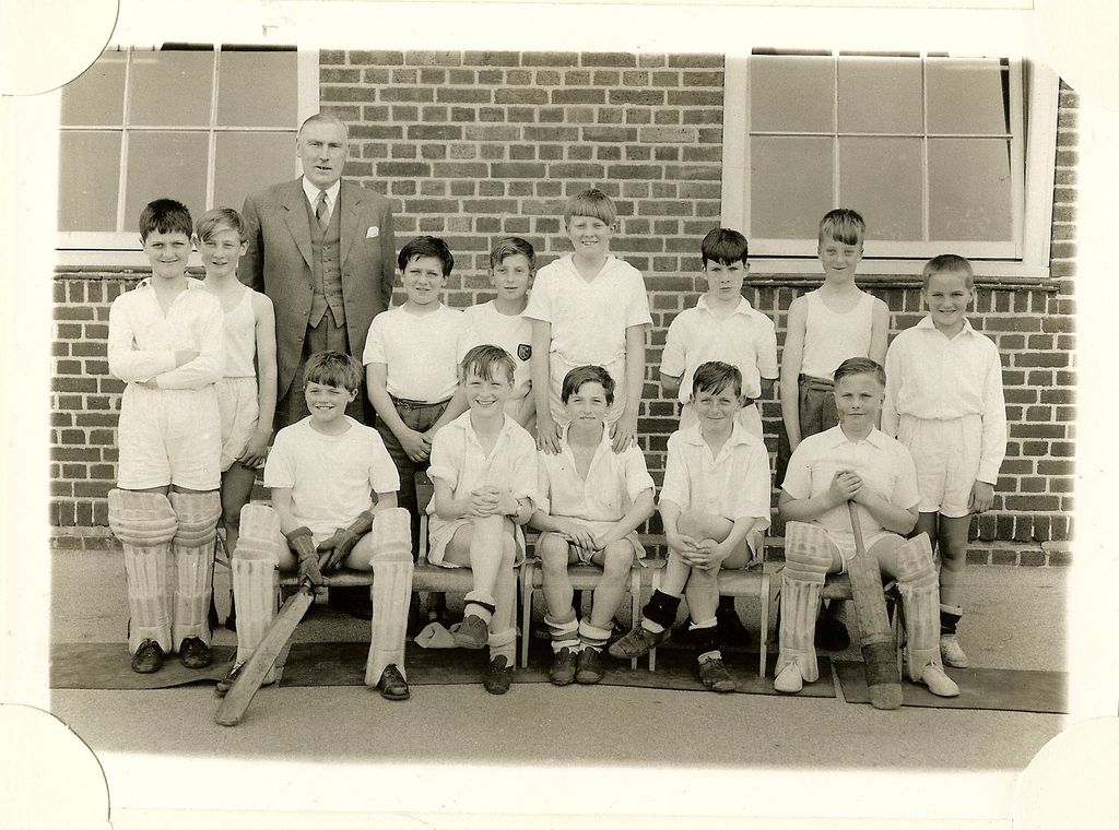 School cricket team of about thirteen boys in whites, two wearing pads and holding bats, with an older man in a three-piece suit standing behind.
