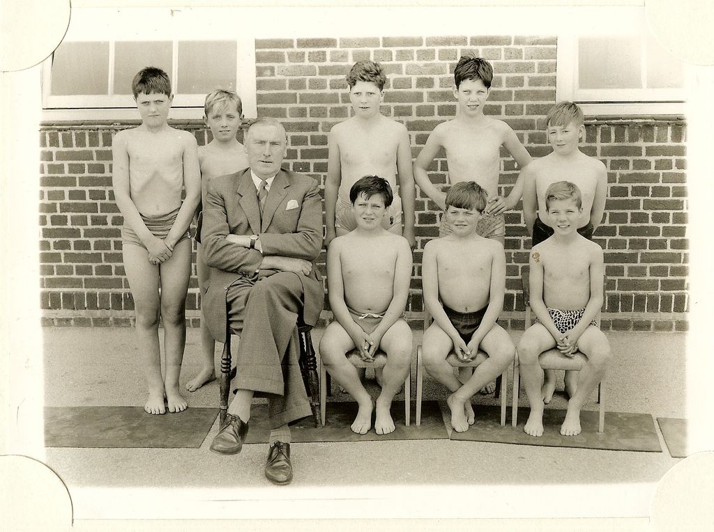 A school team photograph of eight boys in swimming trunks with a headmaster in a suit seated among them, in front of a brick wall.