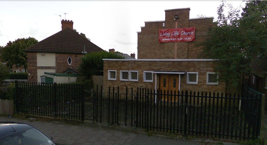 A street view of a brick church hall behind black railings: a single-storey front with a double door and a taller gabled hall behind, hung with a red banner reading “Living Life Church – Sundays at 10.30–12.00”, beside a pair of semi-detached houses. “© 2009 Google” appears faintly on the image.