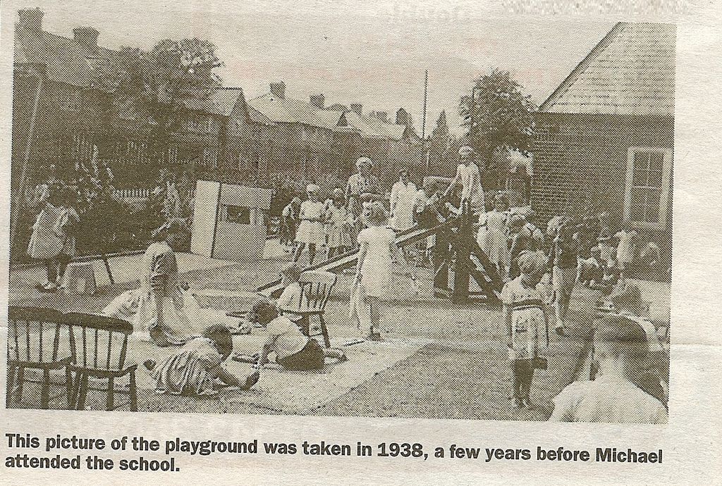 A newspaper photograph of young children playing in a school playground with a seesaw, a small wooden playhouse and chairs, watched by teachers, with houses behind. The printed caption reads “This picture of the playground was taken in 1938, a few years before Michael attended the school.”