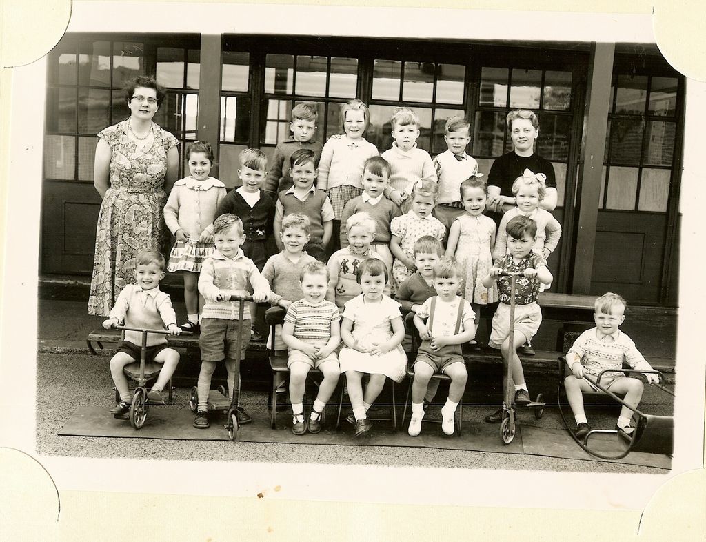 About thirty very young children in four rows with two women teachers, several sitting on tricycles, scooters and a toy car at the front, in front of glazed classroom doors.