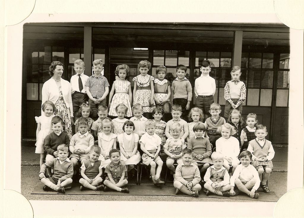 Class of about forty infants in four rows, a woman teacher in a cardigan standing at the left, the front row sitting cross-legged on a mat.
