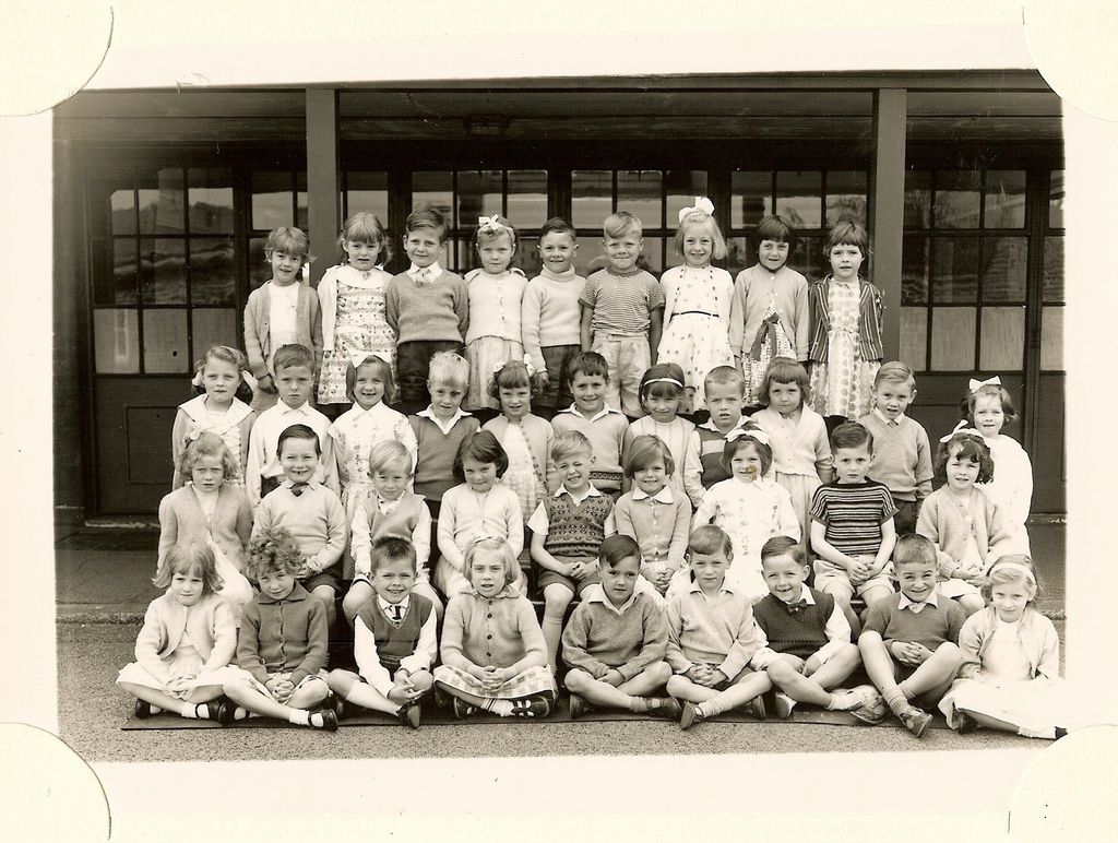 Class of about forty-five infants in four rows, the front row sitting cross-legged, in front of glazed classroom doors; no teacher appears.
