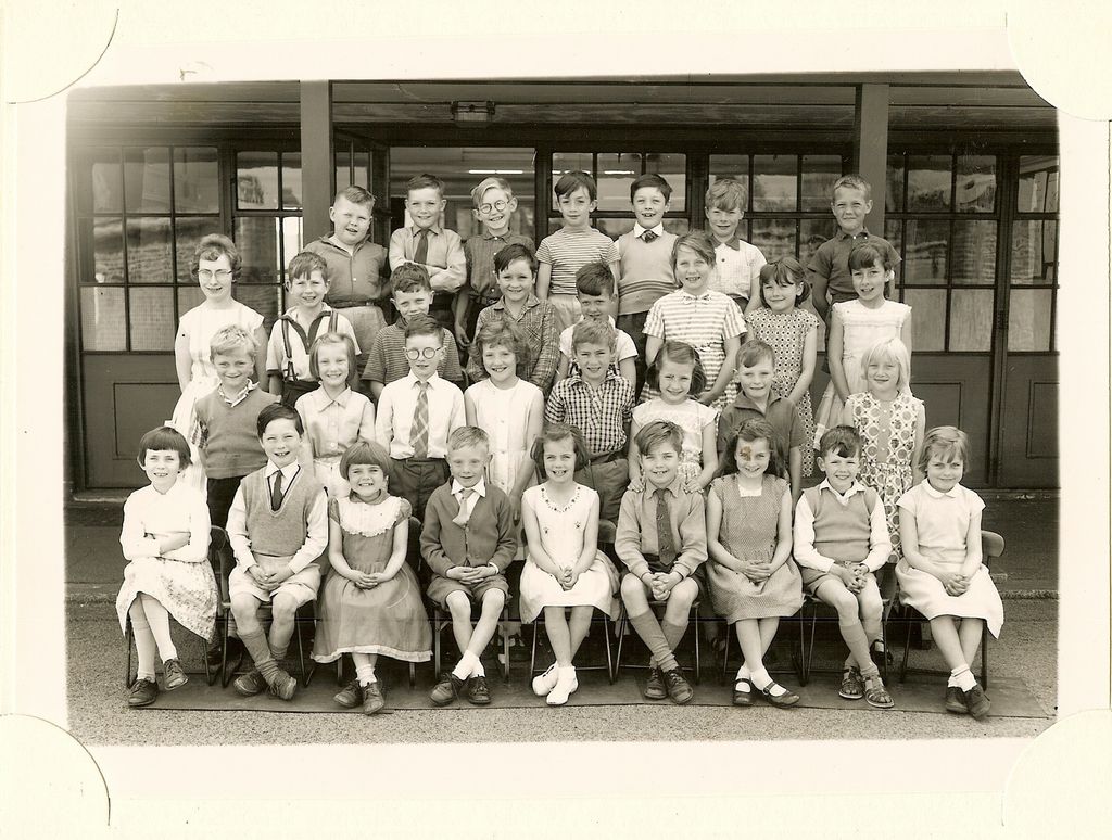 Class of about forty children in four rows, a woman teacher in glasses standing at the left, in front of a veranda with glazed doors.