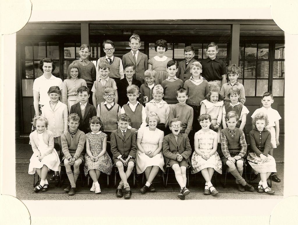 Class of about forty junior-school children in four rows, a woman teacher in a white blouse standing at the left, in front of glazed classroom doors.