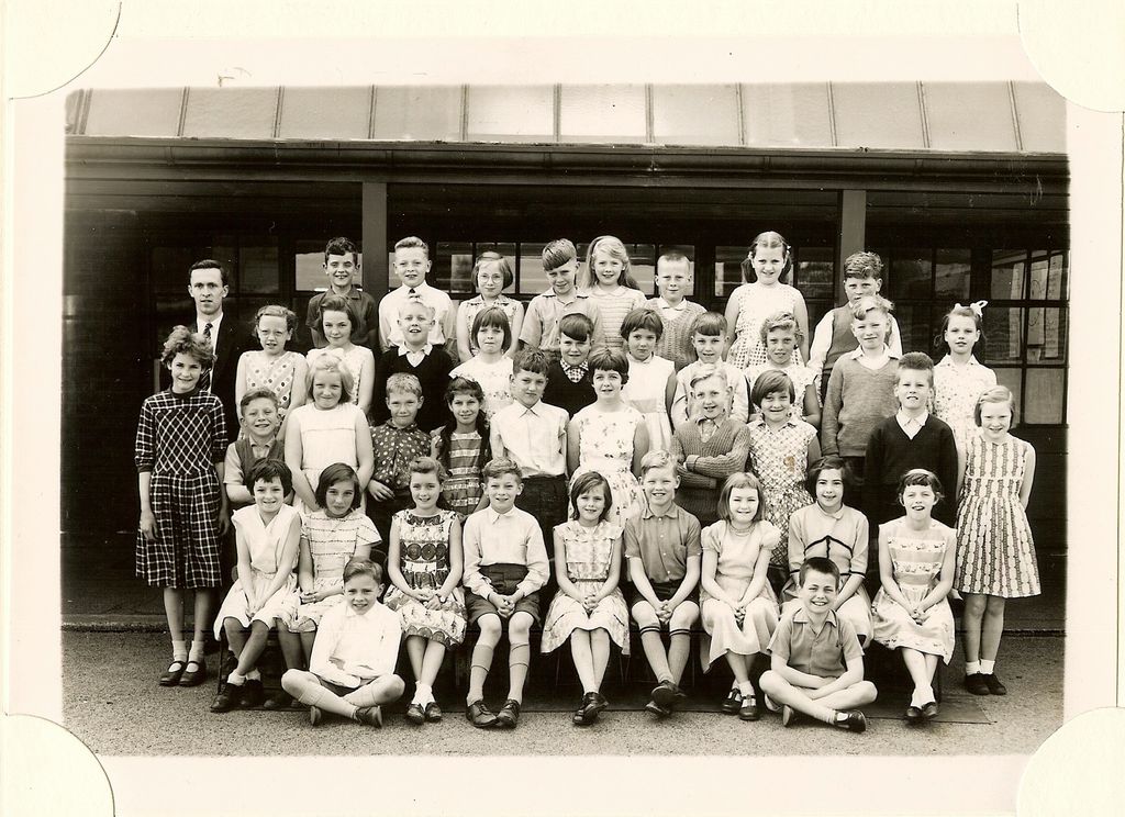 Large class of about fifty junior-school children in six rows under a veranda canopy, a young male teacher standing at the back left.