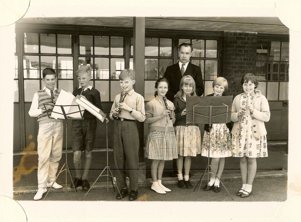 Seven children playing recorders at music stands, with a male teacher in a dark suit standing behind them, in front of glazed classroom doors.