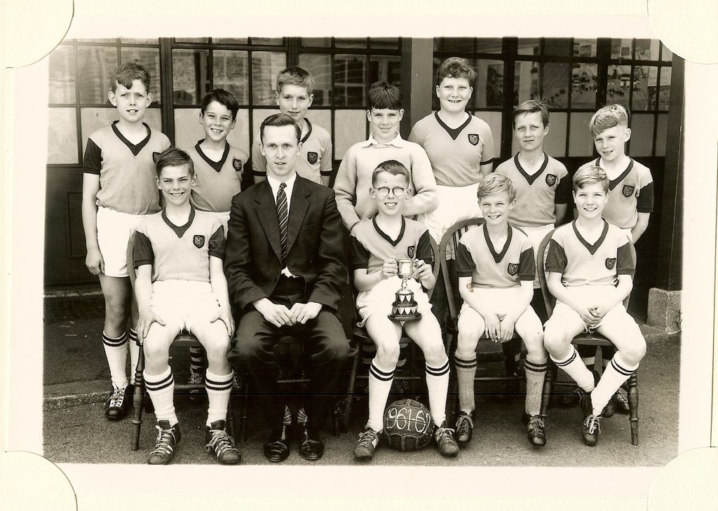 School football team of eleven boys in dark shirts with light collars and hooped socks, one holding a trophy cup, with a young male teacher in a suit; the ball is marked “1961-62”.