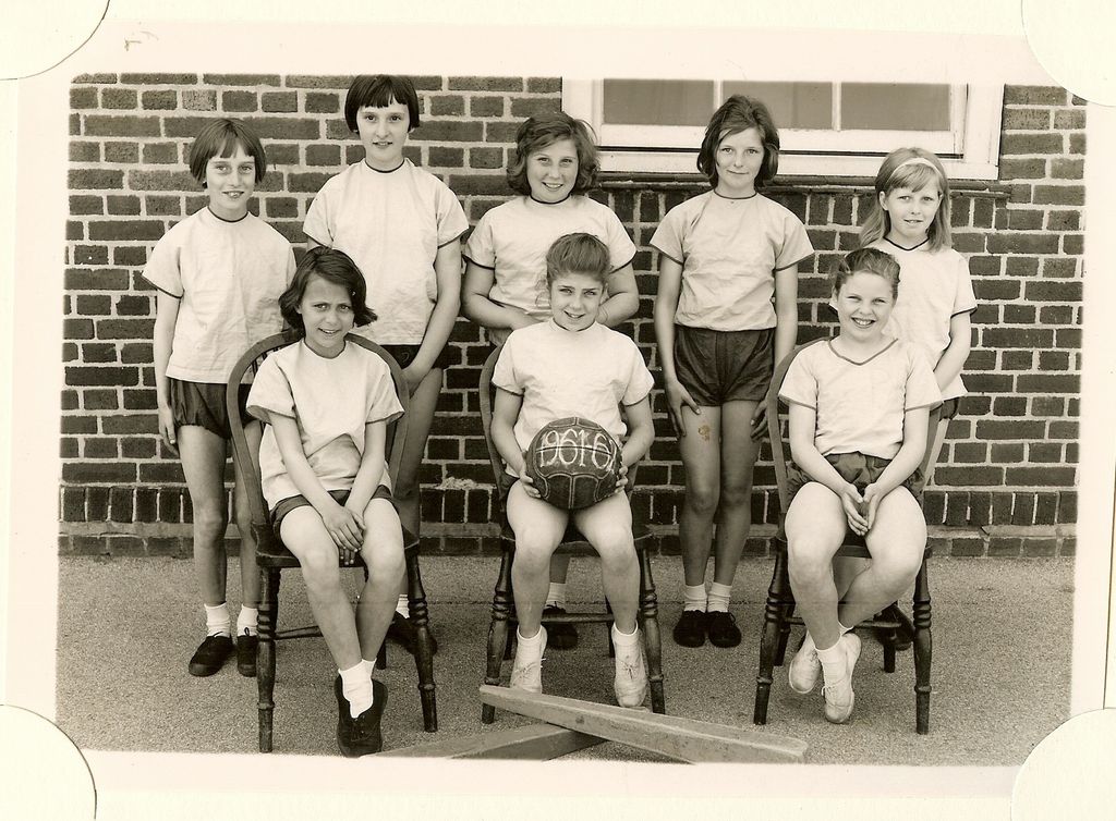 Girls’ sports team of eight girls in light shirts and dark shorts in front of a brick wall, one holding a ball marked “1961-62”, with a bat on the ground in front.