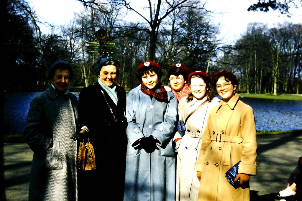 Colour slide of two older women teachers in winter coats and four girls in red berets and long coats standing beside a lake in the parkland at Keukenhof, a signpost behind them.