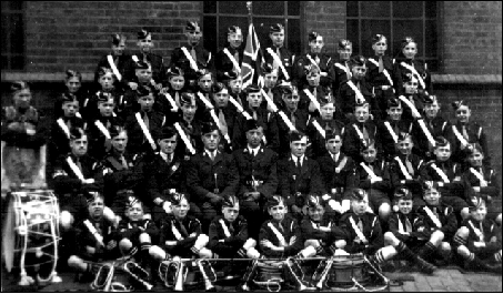 A small photograph of about sixty boys and officers of the Boys’ Brigade in caps and cross-belts, in rows in front of a brick building, with drums, bugles and a flag.