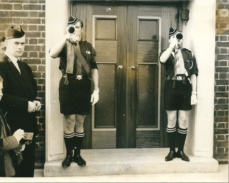 Two Boys’ Brigade buglers in uniform sound bugles either side of the panelled double doors of a new church, while an officer in a glengarry cap and onlookers watch at the left.