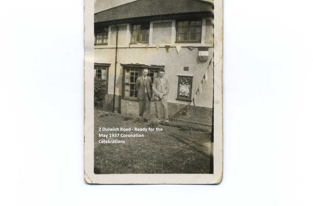 A small worn snapshot of two men in suits standing in front of a rendered house decorated with bunting and a framed royal portrait in the window, with a lawn in the foreground; a caption is printed on the image.