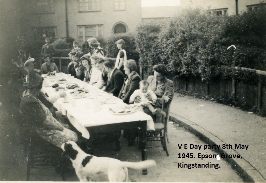 Neighbours, including women holding babies, sit along a long table laid with a white cloth in the curve of a grove, with a black-and-white dog in the foreground; a caption is printed on the image.