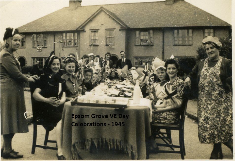 Women and girls in party hats, some holding Union flags, sit at a table in the road with jugs and plates, two older women in aprons standing at either end, in front of rendered semi-detached houses with flags in the windows.