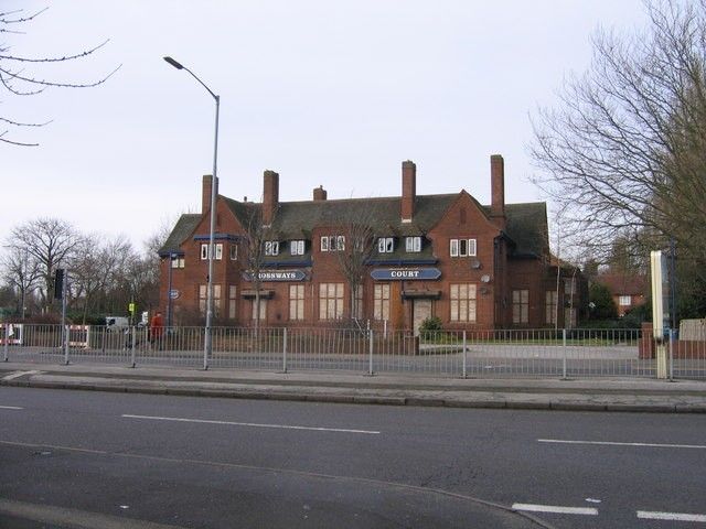 Large brick former pub with tall chimneys and gables, signed 'Crossways Court', standing empty behind railings at a crossroads