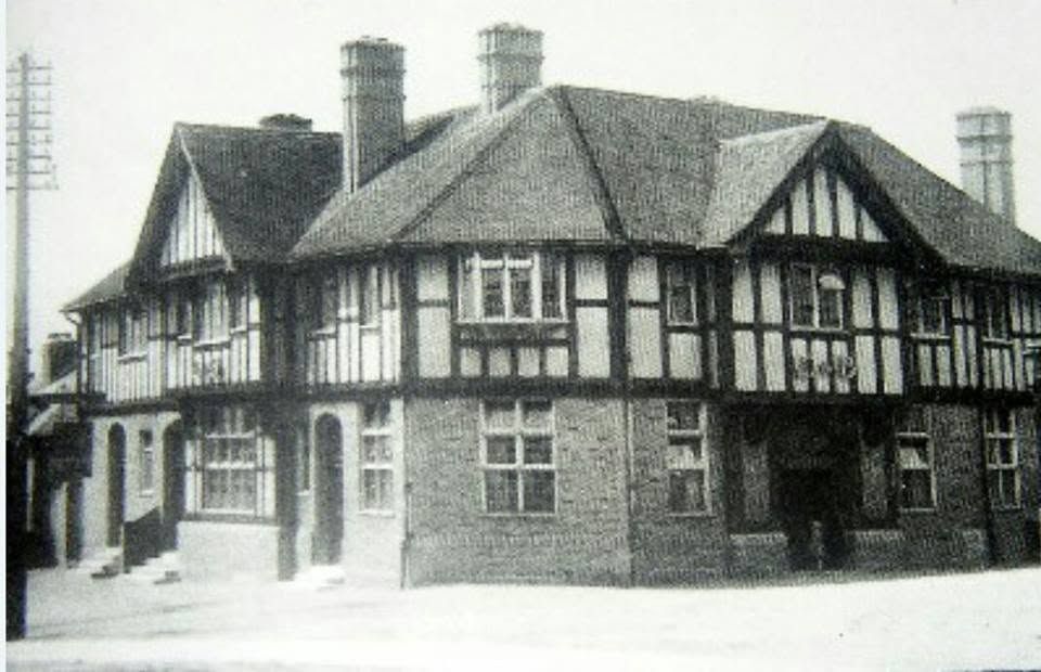 A large mock-Tudor corner building with black-and-white half-timbering, jettied upper floor, tall brick chimneys and a telegraph pole