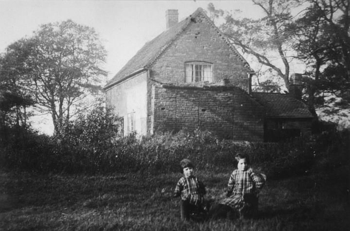 An old brick cottage among trees, with two small children sitting on the grass in front.