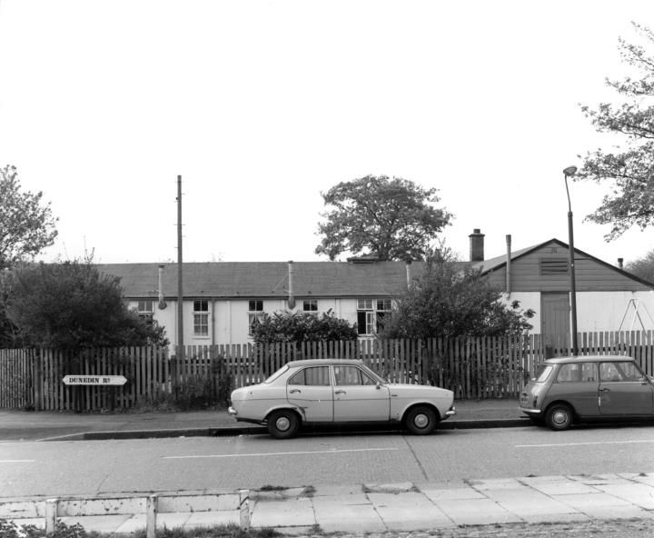 A long single-storey nursery building behind a paling fence on Dunedin Road, with 1970s cars parked outside.