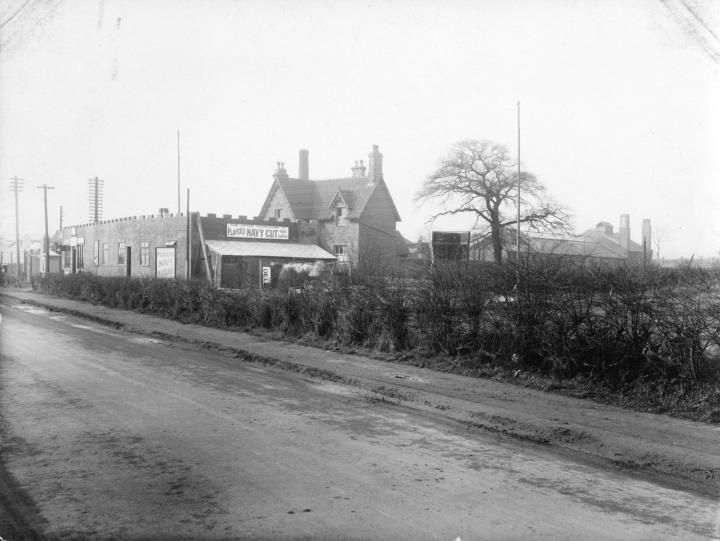 A muddy road with a hedge in the foreground and, behind it, a house and a single-storey shop with advertising signs.