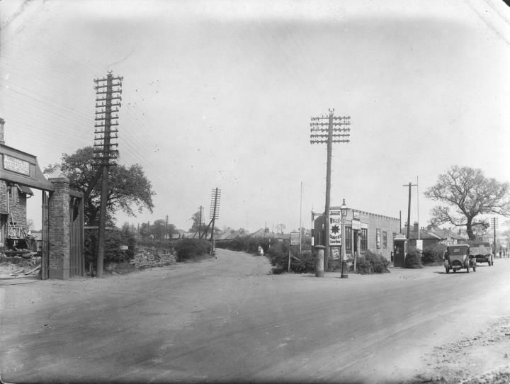 A road junction with telegraph poles, a brick gatepost, a petrol station with enamel signs and 1930s cars.
