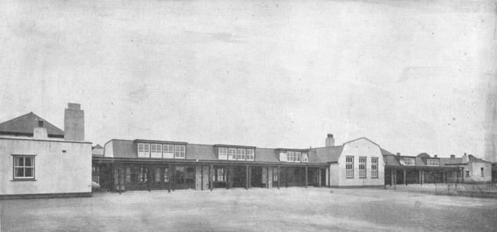 A long low school building with a verandah along its front, facing a wide open playground.