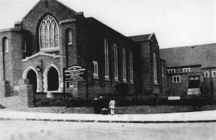 A brick Gothic-style church with arched entrance doors and a large pointed window, with a notice board outside.