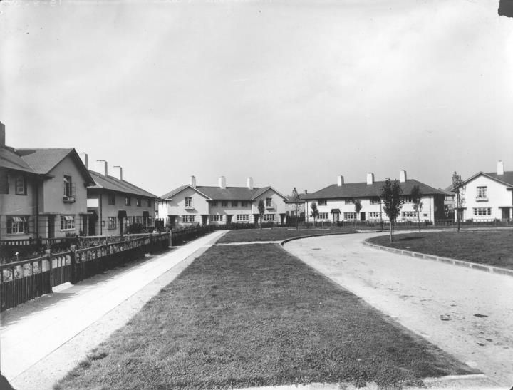 A long grass strip between two paths leading towards groups of white rendered houses, with low fencing along the front gardens.
